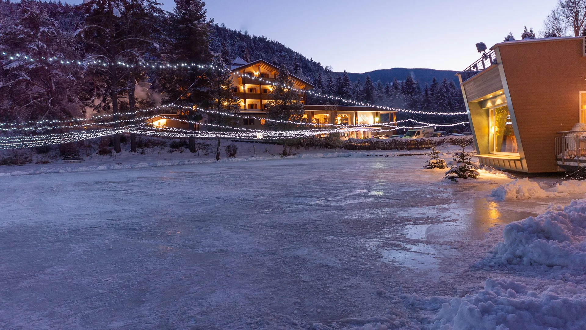 Natureislaufplatz Issinger Weiher Verschneite Winterlandschaft mit beleuchtetem Hotel und zugefrorenem See