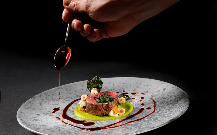Hand träufelt Sauce auf kunstvoll angerichtetes Gourmetgericht auf grauem Teller