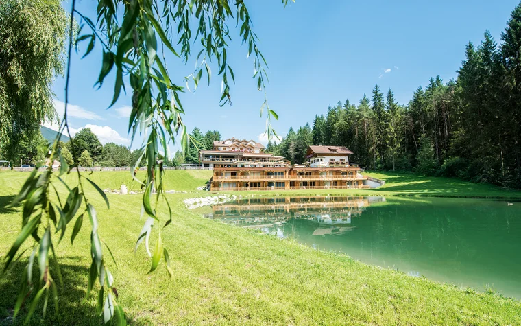 Hotelgebäude am Seeufer mit grüner Wiese und Wald im Hintergrund