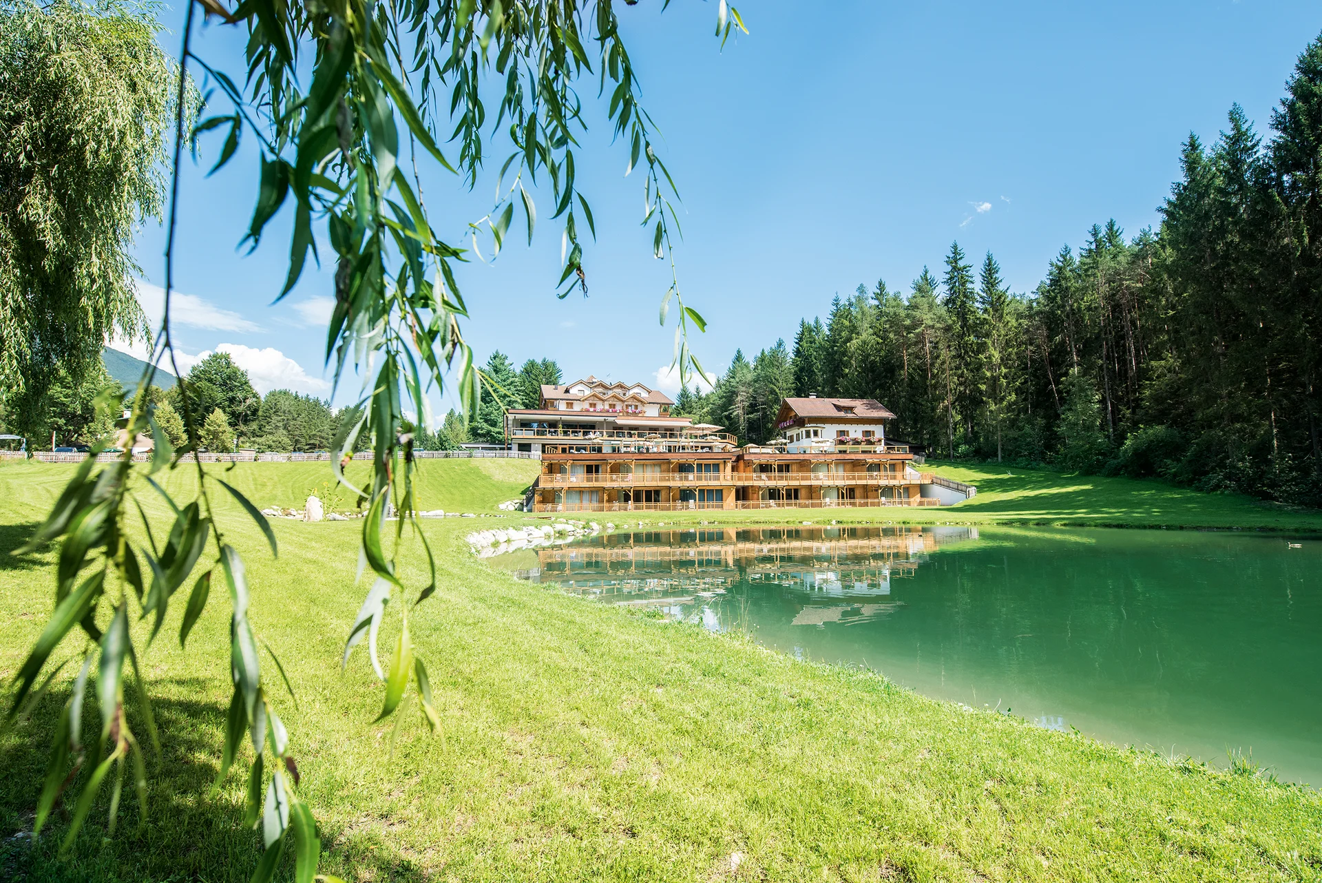 Natureislaufplatz Issinger Weiher Hotelgebäude am Seeufer mit grüner Wiese und Wald im Hintergrund