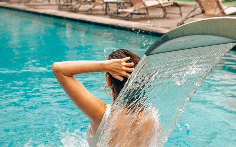 Frau entspannt sich unter Wasserfall im Pool mit Liegestühlen im Hintergrund