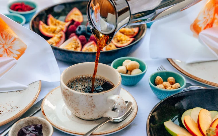 Kaffee wird in eine Tasse gegossen, umgeben von Früchten und Snacks auf einem gedeckten Tisch