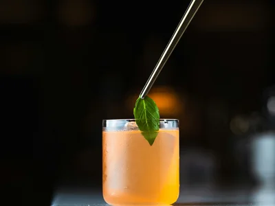 Hand platziert Minzblatt mit Pinzette auf orangefarbenem Cocktail im Glas