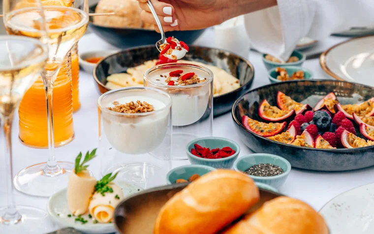 Frühstückstisch mit Joghurt, Obst, Brötchen und Getränken auf weißer Tischdecke
